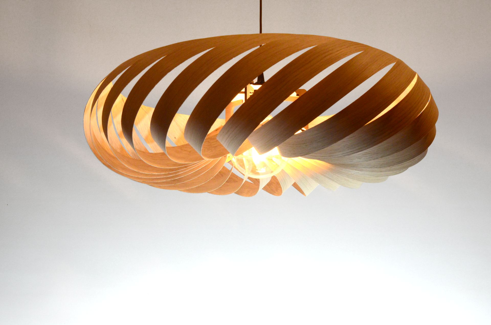 Suspension Vortex M en bois de chêne, luminaire sculptural aux lames courbées diffusant une lumière chaude.